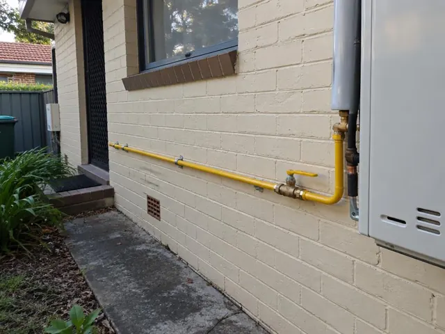 Yellow exterior gas pipework on a brick house wall beside a window and meter box in Ermington